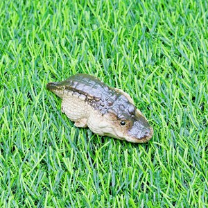 May include: A small alligator figurine with a textured, dark green and brown body, set on bright green artificial grass. The figurine has a detailed face and a light-coloured underbelly. A decorative item.