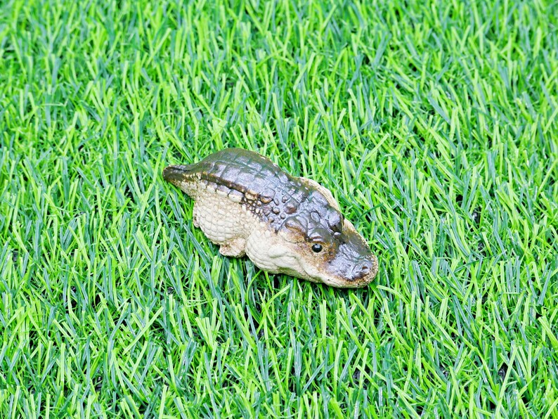 May include: A small alligator figurine with a textured, dark green and brown body, set on bright green artificial grass. The figurine has a detailed face and a light-coloured underbelly. A decorative item.