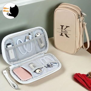 Op de afbeelding: Een witte reistas met rits met een grijs interieur en meerdere vakken om elektronica en accessoires te organiseren. De tas is open en toont een telefoon, oplaadkabels en andere kleine items. De tas heeft een witte band en een beige ritsvak met de naam "Kally" erop.