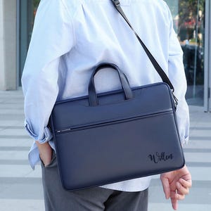 May include: A navy blue laptop bag with a handle and a shoulder strap. The bag has a zipper pocket and the word "Willen" is written on the front. The bag is being carried over the shoulder.