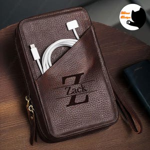 May include: A brown leather tech organiser with a zip closure and a wrist strap. The organiser has a pocket holding a white charging cable. The name "Zack" is printed on the front, with the letter "Z" below it.