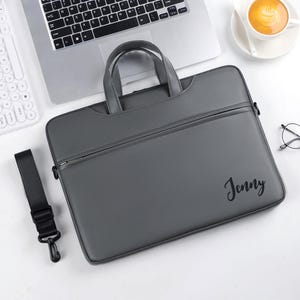 May include: Gray laptop bag with a handle and a shoulder strap. The bag has a zipper closure and the name "Jenny" printed on the front. A laptop, keyboard, and a cup of coffee are in the background.