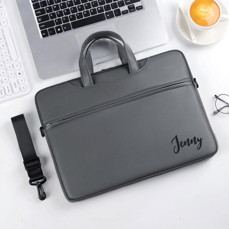 May include: Gray laptop bag with a handle and a shoulder strap. The bag has a zipper closure and the name "Jenny" printed on the front. A laptop, keyboard, and a cup of coffee are in the background.