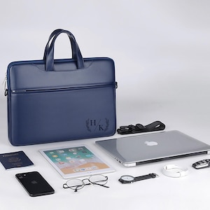 May include: A navy blue leather briefcase with a handle, zipper, and a front pocket. The briefcase has the initials "H/K" with a laurel wreath design. A laptop, tablet, phone, glasses, watch, passport, and other accessories are arranged around the briefcase.