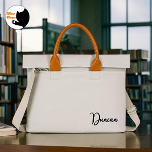 May include: A white tote bag with a brown handle and shoulder strap. The bag has the name "Duncan" printed in black script. The bag has a zipper closure and a flap that folds over the top.