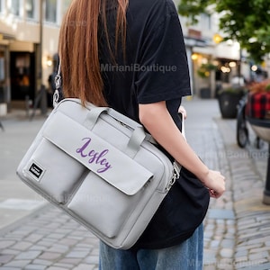 May include: A gray laptop bag with a shoulder strap and a handle. The bag has a flap closure and two front pockets. The name "Lesley" is written in purple script on the front of the bag. The bag is being carried outdoors.