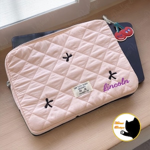 May include: A pale pink quilted laptop sleeve with a diamond pattern and black cross-shaped details. The name "Lincoln" is written in purple script. A cherry charm hangs from the zipper. A small tag with text is visible.