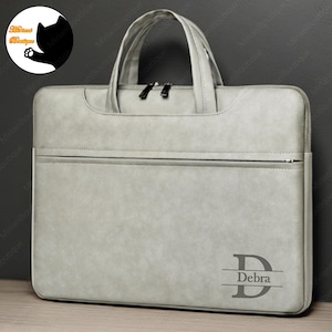 May include: A light gray laptop bag with a zipper closure and a top handle. The bag has a personalized name, "Debra", engraved on the front.