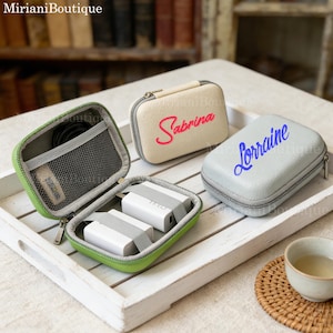 May include: Several personalised travel cases in different colours. One case is open, showing white chargers and a mesh pocket. Two closed cases are visible: one beige with "Sabrina" in pink, and a grey one with "Lorraine" in blue.