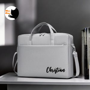 Op de afbeelding: Een grijze laptoptas met een zwarte rits en een zwarte band. De tas heeft de naam "Christian" erop geschreven in zwarte cursieve letters.