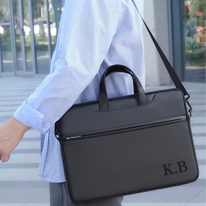 May include: A black briefcase with a shoulder strap and a top handle. The briefcase has a zipper closure and the initials "K.B" printed on the lower right corner. The person in the image is wearing a light blue button-down shirt.