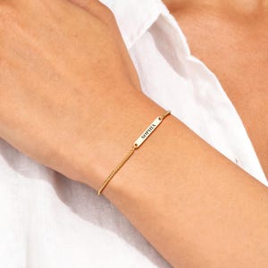 Pode incluir: Uma pulseira dourada com uma corrente delicada e uma barra retangular gravada com o nome "SOPHIA". A pulseira é usada no pulso. O fundo é branco.