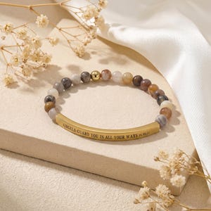 May include: A beaded bracelet with a gold-colored curved bar. The bracelet features a variety of earth-toned beads, including brown, gray, and cream colors. The gold bar is inscribed with the words "ANGELS GUARD YOU IN ALL YOUR WAYS PS. 91:11".