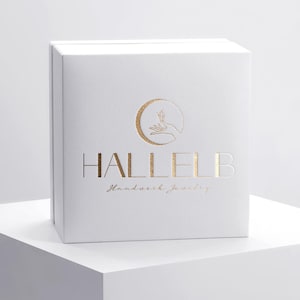 May include: A white box with gold lettering and a logo featuring a crescent moon and a hand. The box reads "HALLELB Handwork Jewelry". The box is sitting on a white surface.