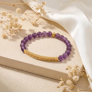 May include: A beaded bracelet with amethyst stones and a gold-colored bar. The bar is engraved with the words "ALWAYS MY MOTHER, FOREVER MY FRIEND". The bracelet is displayed on a white surface with dried flowers.