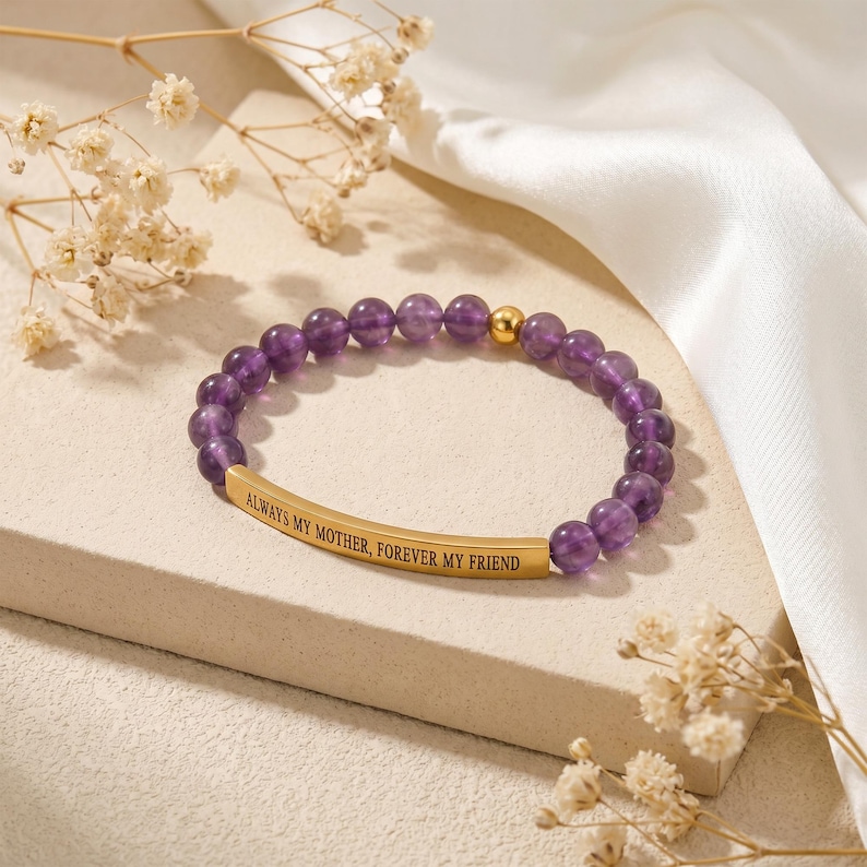 May include: A beaded bracelet with amethyst stones and a gold-colored bar. The bar is engraved with the words "ALWAYS MY MOTHER, FOREVER MY FRIEND". The bracelet is displayed on a white surface with dried flowers.