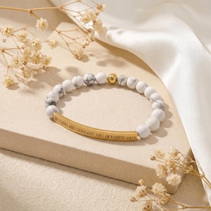 May include: White and gray beaded bracelet with a gold-colored bar inscribed with the words "BE STRONG AND COURAGEOUS FOR I AM WITH YOU JOSH 1:9". The bracelet also features a gold-colored bead.