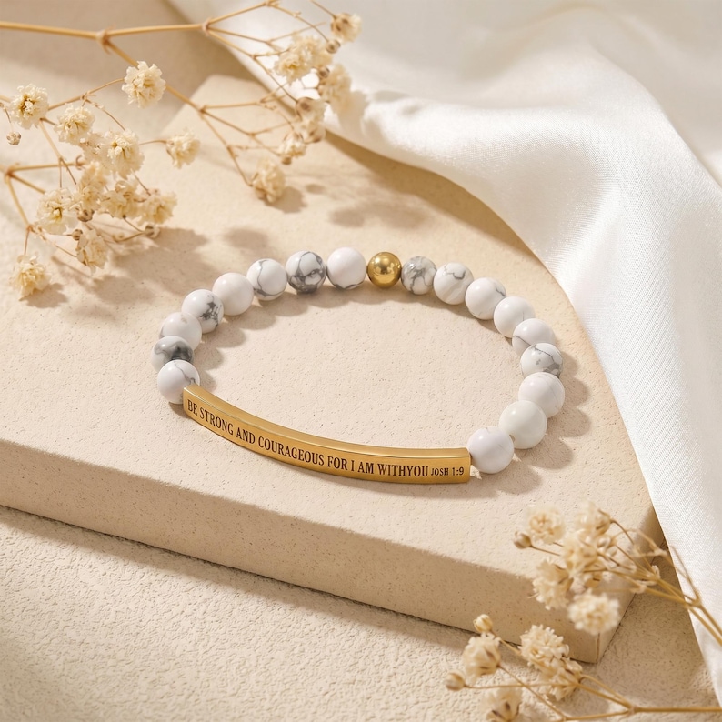 May include: White and gray beaded bracelet with a gold-colored bar inscribed with the words "BE STRONG AND COURAGEOUS FOR I AM WITH YOU JOSH 1:9". The bracelet also features a gold-colored bead.