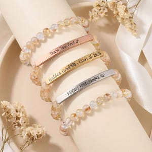 May include: Four beaded bracelets with engraved metal bars. The bracelets feature light-colored, round beads. The metal bars are engraved with the phrases: "Thank You, Prof", "Golden Grad - Class of 2025", and "FRIENDS FOREVERMORE".