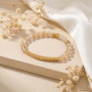 May include: A beaded bracelet with golden and clear stones. A gold bar with the inscription "Golden Grad - Class of 2025" is centered on the bracelet. The bracelet is displayed on a light beige surface with dried flowers and a white satin cloth.