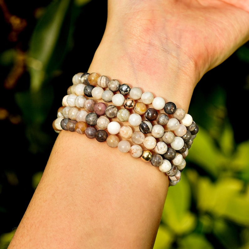 May include: A stack of beaded bracelets in neutral tones of white, gray, brown, and pink. The bracelets are made of small, round beads and accented with gold-colored beads. The bracelets are stacked on a wrist.