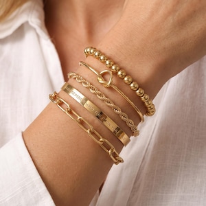 Peut inclure: Un ensemble de cinq bracelets dorés comprenant un rang de perles, un bracelet fin avec un nœud, une chaîne torsadée, un jonc plat gravé avec le texte REMEMBER WHO THE FUCK YOU ARE et un bracelet à maillons ovales épais.