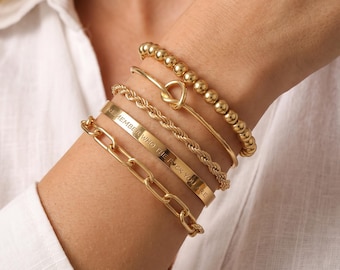 May include: A set of five gold-toned bracelets including a beaded strand, a thin band with a knot detail, a twisted rope chain, a flat cuff engraved with the text REMEMBER WHO THE FUCK YOU ARE, and a chunky oval link chain bracelet.