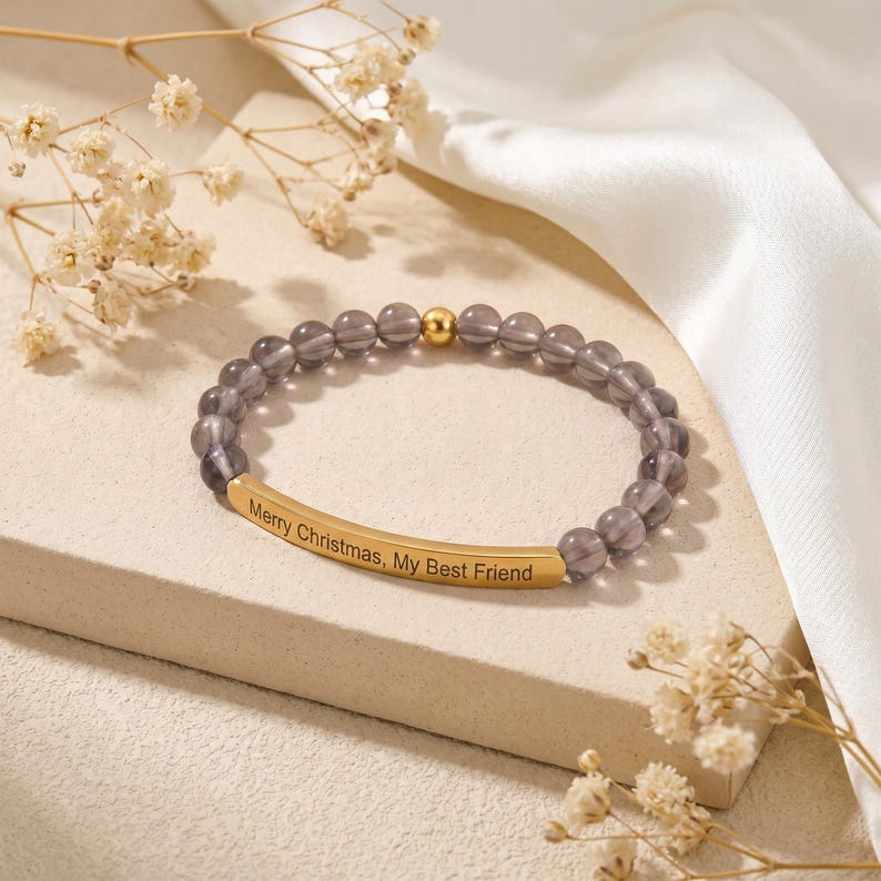 May include: A beaded bracelet with gray beads and a gold-colored bar engraved with "Merry Christmas, My Best Friend." The bracelet also features a gold-colored bead. The bracelet is displayed on a light-colored surface with dried flowers.