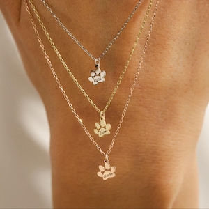 Puede incluir: Tres collares de oro con dijes en forma de huella de pata. Los dijes están grabados con los nombres "Luna", "Jack" y "Charlie".
