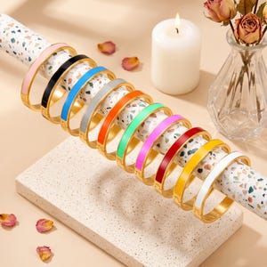 Könnte beinhalten: Eine Kollektion von zehn aufklappbaren Armreifen mit einer goldfarbenen Metallbasis und einer glatten, farbigen Emaille-Einlage. Die Armreifen sind in einer Reihe angeordnet und zeigen Farben wie Rosa, Schwarz, Blau, Silber, Orange, Grün, Violett, Rot, Gelb und Weiß.