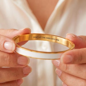 Könnte beinhalten: Ein goldfarbener Metallarmreif mit einer glatten weißen Emaileinlage auf der Außenseite. Die Innenseite des Armreifs ist mit dem schwarzen Text DON'T LET THE HARD DAYS WIN in einer klaren, serifenlosen Großbuchstabenschrift graviert.
