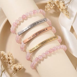 May include: Four beaded bracelets with pink gemstones. Each bracelet features a metal bar with an engraved message: "I LOVE YOU TO THE MOON AND BACK", "YOU ARE MY SUNSHINE", and "BEAUTIFUL GIRL, YOU WERE MADE TO DO GREAT THINGS."