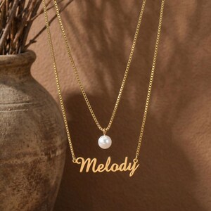 Può includere: Una collana dorata a due strati con un delicato design a catena. Lo strato superiore presenta un ciondolo di perla bianca, mentre lo strato inferiore riporta il nome "Melody" in corsivo. Lo sfondo è di un tono caldo e neutro.