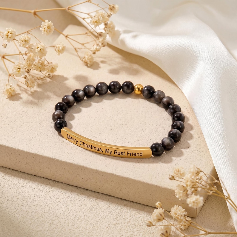 May include: A beaded bracelet with dark gray and gold-colored beads. A gold-colored bar is engraved with the words "Merry Christmas, My Best Friend". The bracelet is displayed on a light-colored surface with dried flowers and a white satin cloth in the background.