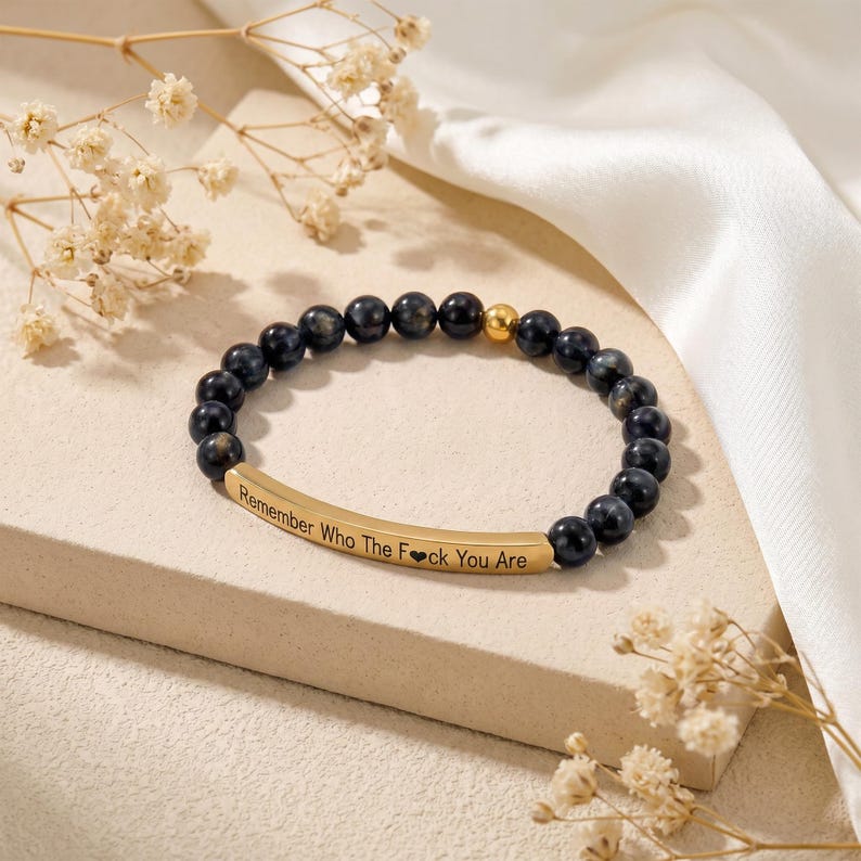 May include: A beaded bracelet with dark blue spherical beads and a gold-colored accent bead. A gold-colored curved bar with the engraved text "Remember Who The F*ck You Are" is centered on the bracelet. The bracelet is displayed on a light-colored surface.