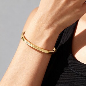 May include: A gold-colored, rectangular bangle bracelet. The bracelet has the engraved text "BEAUTIFUL GIRL, YOU WERE MADE TO DO GREAT THINGS". The bracelet is worn on a person's wrist.