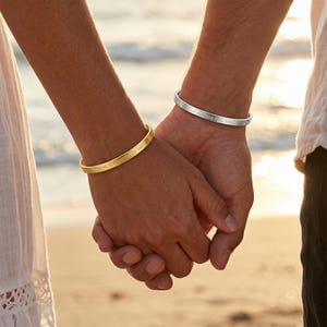 May include: Two hands holding each other, each wearing a bracelet. One bracelet is gold-colored, the other is silver-colored. The gold bracelet has text engraved on it. The background is a beach scene with the sun setting.