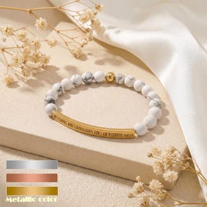 May include: A bracelet with white and grey marble-effect beads, a gold-coloured bead, and a gold-coloured curved bar inscribed with "BE STRONG AND COURAGEOUS FOR I AM WITH YOU JOSH 1:9". The bracelet is displayed on a neutral surface.