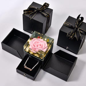 May include: Black gift boxes with textured surfaces and black ribbons with gold text. One box contains a preserved pink rose in a glass case. Another box holds a necklace with the name "Tammy".