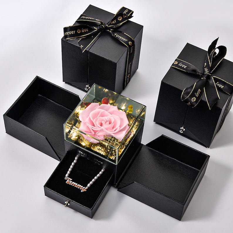 May include: Black gift boxes with textured surfaces and black ribbons with gold text. One box contains a preserved pink rose in a glass case. Another box holds a necklace with the name "Tammy".