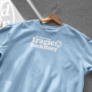 Op de afbeelding: Een lichtblauwe sweatshirt met de witte tekst "ask about my tragic backstory". De sweatshirt hangt aan een houten hanger tegen een grijze achtergrond. Een wit logo met de woorden "Crooked Hollow" staat in de linkerbovenhoek.