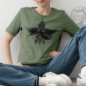 Könnte beinhalten: Ein grünes T-Shirt mit einem schwarzen Motiv eines Vogels mit ausgebreiteten Flügeln, einem Herzen und einer Krone. Der Text "Crooked Hollow" ist im Hintergrund sichtbar.
