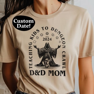 Puede incluir: Camiseta beige con un gráfico negro que presenta una figura con túnica, un niño y dados. El texto dice "TEACHING KIDS TO DUNGEON CRAWL" y "D&D MOM" con el año "2024". Un óvalo negro con "Custom Date!" está en el hombro.