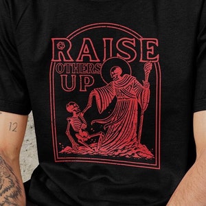 Könnte beinhalten: Schwarzes T-Shirt mit einem roten Grafikdruck eines Skeletts, das einen Stab hält, und dem Text "RAISE OTHERS UP".