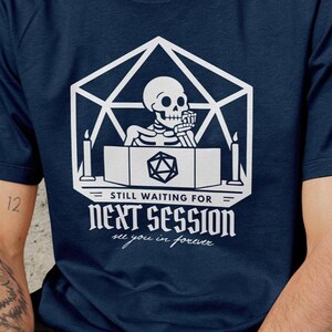 Puede incluir: Camiseta azul marino con un diseño blanco. El diseño incluye un esqueleto, una forma geométrica, velas y el texto "STILL WAITING FOR NEXT SESSION all you in forever". La camiseta tiene cuello redondo.