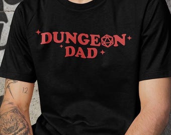 以下が含まれることがあります： 赤い「DUNGEON DAD」の文字とサイコロのグラフィックが特徴の黒いTシャツ。クルーネックのスタイルです。黒いパンツを履いており、腕にタトゥーが入っています。