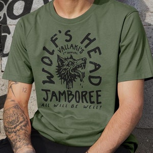 May include: A green t-shirt featuring a black wolf's head graphic and the text "Wolf's Head Vallaki's 1st Annual Jamboree All Will Be Well!"
