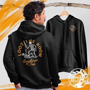 Puede incluir: Sudadera con capucha negra con un gráfico de esqueleto en la espalda, sosteniendo una espada. El texto "LOOT THE ROOM" y "Everything's Free" es dorado. Se muestra una segunda sudadera con capucha en una percha.