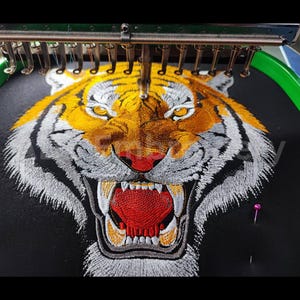 May include: Close-up of an embroidered tiger face with orange, white, and black thread. The tiger's mouth is open, revealing sharp teeth and a red tongue. The embroidery is being created on a black fabric.