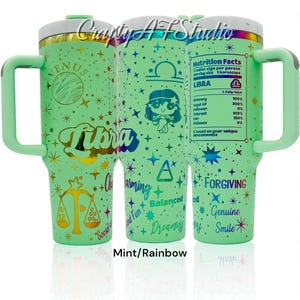 May include: A mint green tumbler with a handle, featuring a Libra zodiac design. The tumbler has a white lid and is decorated with stars, the word "Libra", and the words "Charming", "Balanced", "Dreamy", "Forgiving", "Genuine", and "Smile".